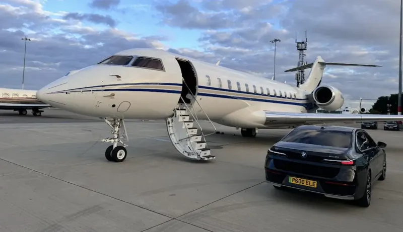 White Gulfstream private jet with open airstair on tarmac beside black BMW car at airport under cloudy sky