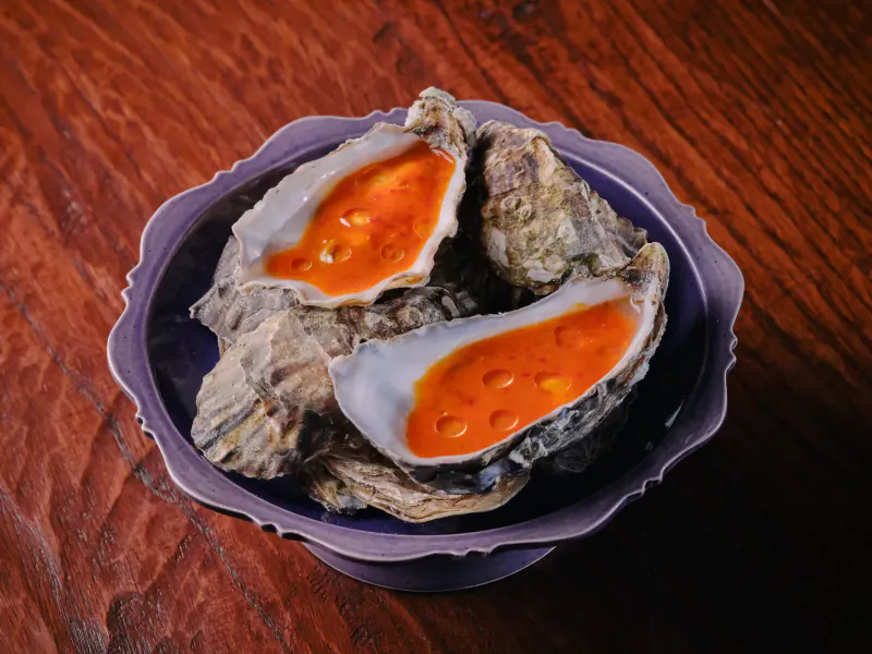Three fresh oysters topped with vibrant orange sauce in a purple dish on wooden table
