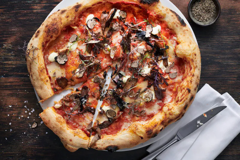 Wood-fired pizza topped with mushrooms, greens, and cheese, sliced on a plate with knife on wooden table