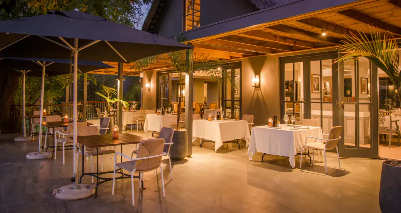 Elegant outdoor terrace dining at Stanley & Livingstone Boutique Hotel, Victoria Falls, with white-clothed tables under umbrellas and palms at dusk.