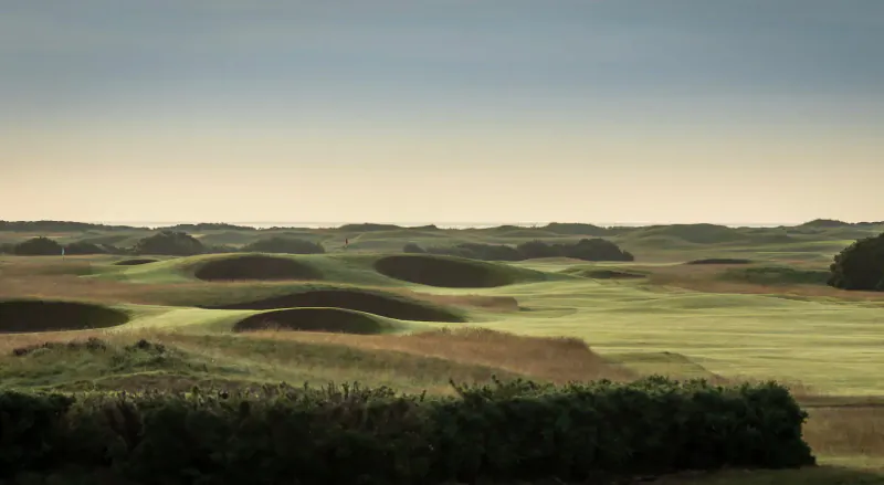 Sweeping view of Carnoustie Golf Links' rolling green fairways and dunes under a hazy sky.