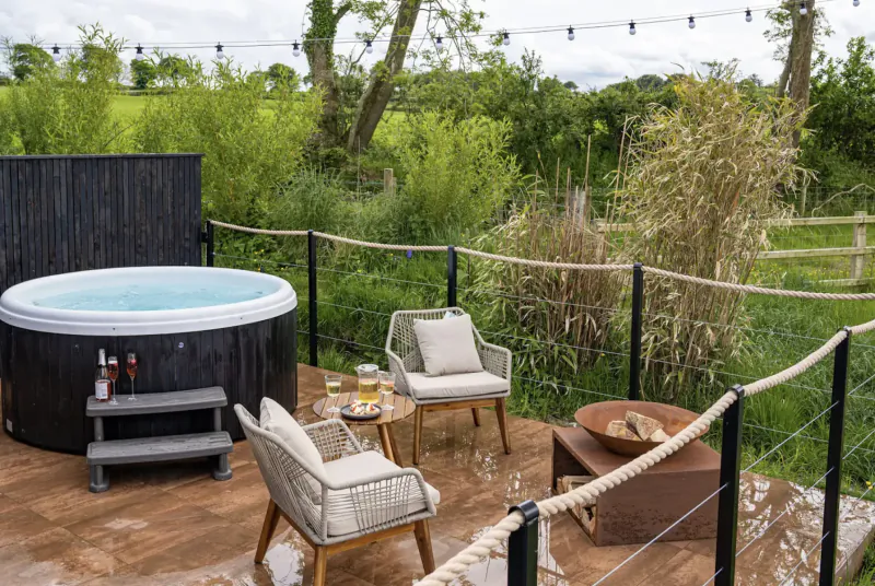 Outdoor hot tub on wooden deck at The Boathouse lodge, with lounge chairs, fire pit, drinks, string lights, and lush green countryside.