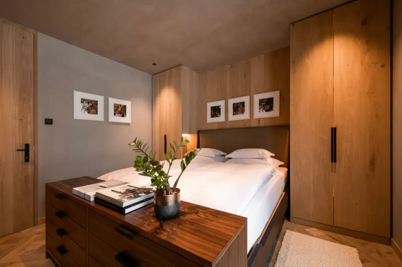 Stylish Austrian chalet bedroom with white duvet bed, wooden wardrobe, dresser with plant, and art on walls.