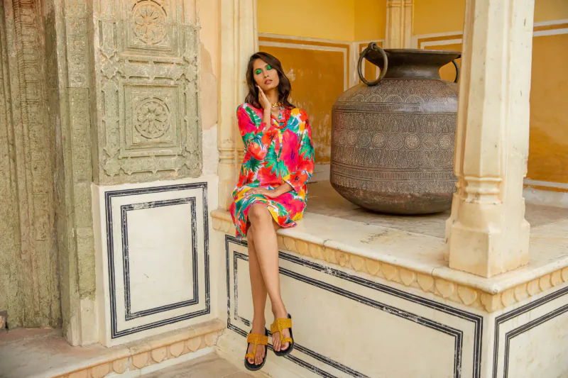 Woman in vibrant floral dress sits on ledge by large pot in ornate yellow Jaipur pavilion