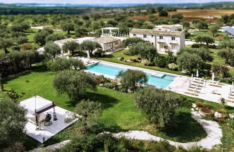 Aerial view of Zahir Country House in Sicily: stone villas, turquoise pool, olive groves, loungers, and pergola amid lush fields.