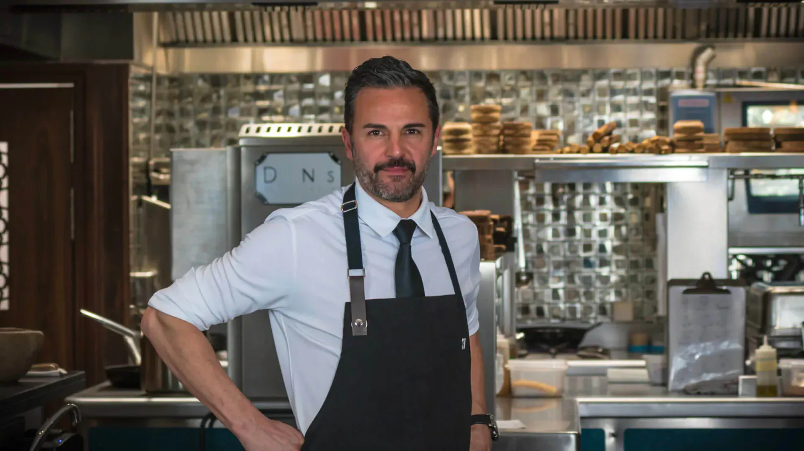 Chef with goatee and salt-and-pepper hair stands confidently in DINS Santi Taura kitchen, wearing white shirt, black tie, and apron amid stainless steel counters and tiles.