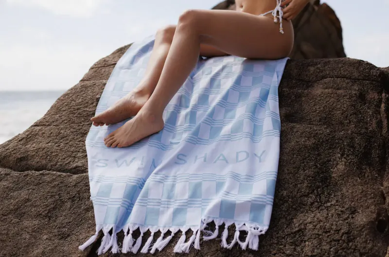 Woman in bikini sits on rock by sea, legs on blue 'Swim Shady' beach towel with fringe.