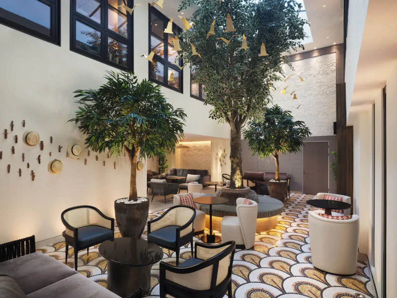 Spacious modern atrium lounge at The Other House with tall potted trees, black chairs, round tables, and geometric tiled floor under skylight.