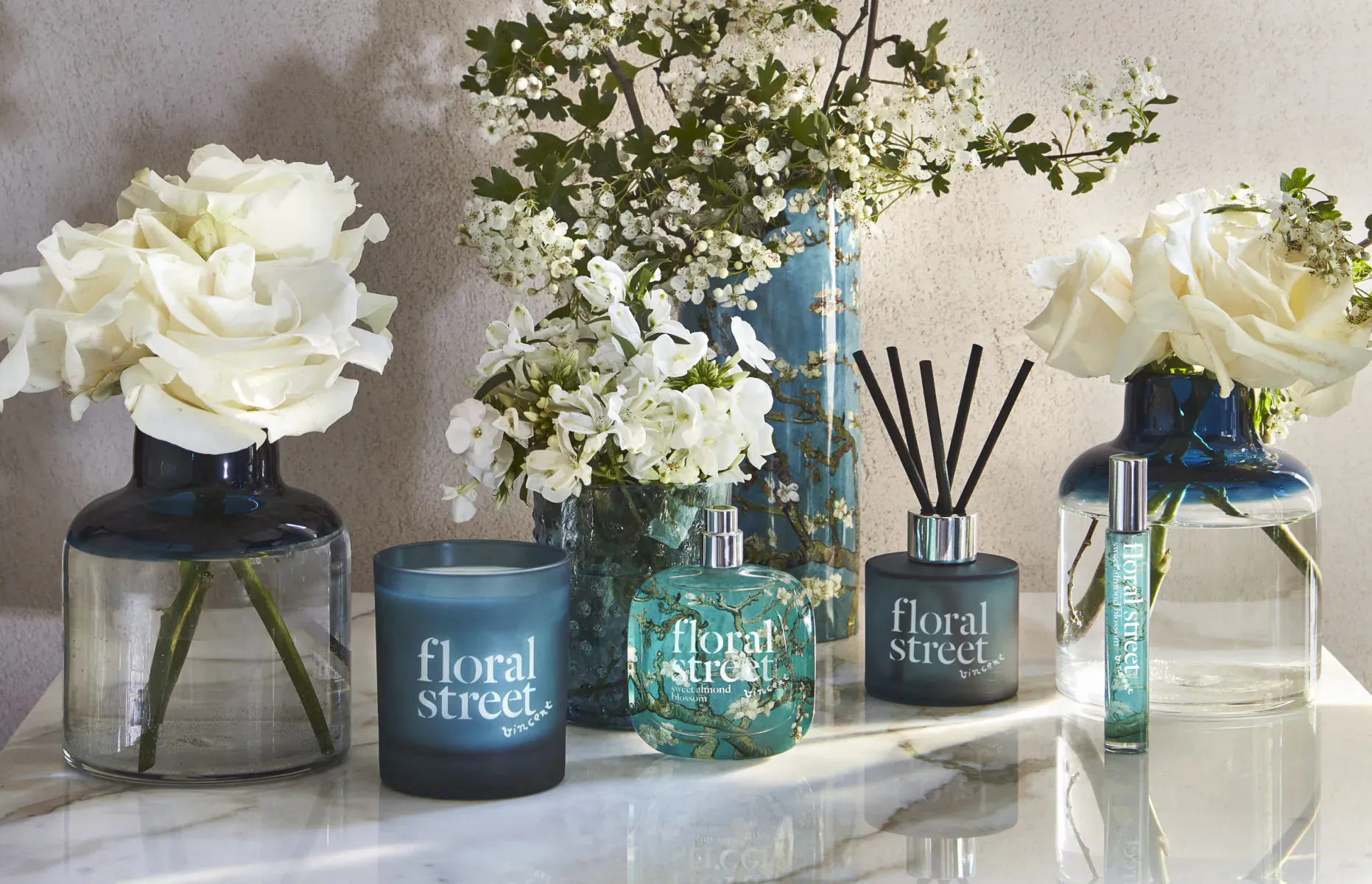 Floral Street sweet almond blossom fragrance collection on table with white roses, branches, blue reed diffuser, candle, and perfume bottle.