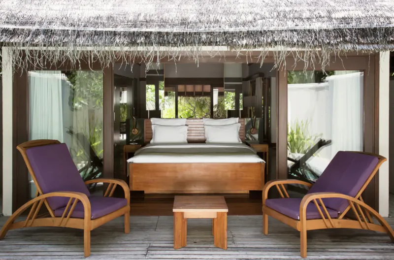 Luxurious open-air bedroom at Coco Bodu Hithi resort with wooden bed, purple armchairs, low table on rug, thatched roof, tropical views.