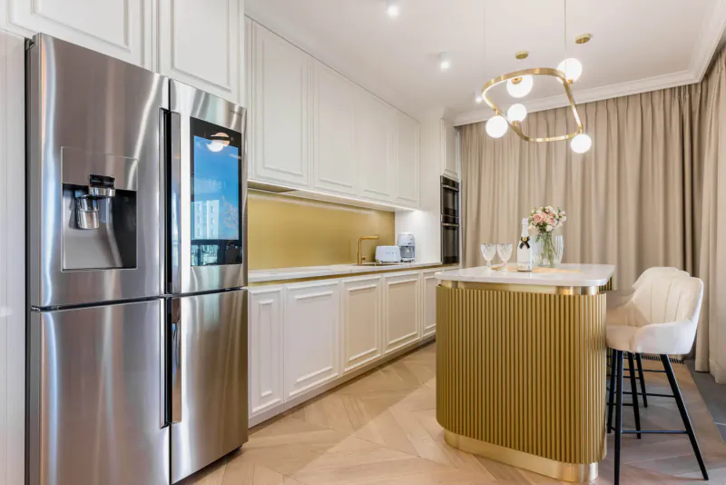 Modern kitchen in luxury apartment with stainless steel fridge, gold-accented island, white cabinets, pendant lights, and flowers.