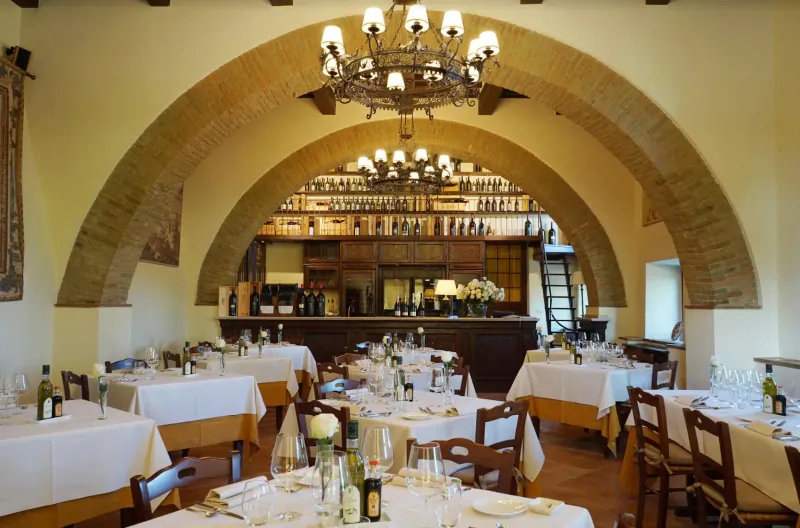 Elegant Tuscan arched dining room with white tablecloths, wine bottles behind wooden bar, chandeliers.