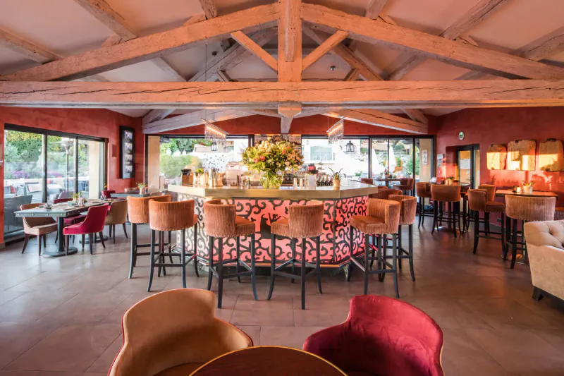 Elegant Hôtel Byblos St Tropez bar with wooden beams, pink central counter, orange stools, flowers, and large windows