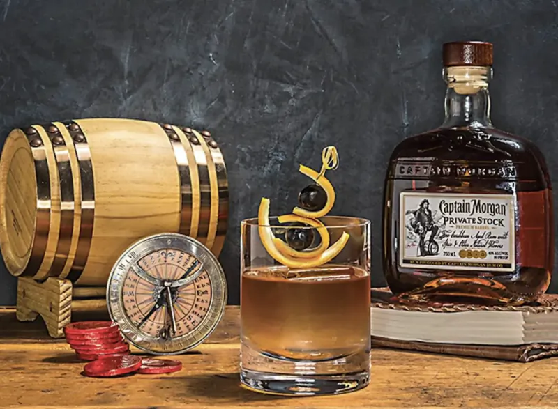 Captain Morgan Private Stock rum old fashioned cocktail with orange twist, beside wooden barrel, compass, and nautical decor.