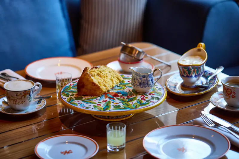 Elegant Italian restaurant table with slice of yellow cake on colorful cake stand, ornate teacups, saucers, milk jug, and glasses.