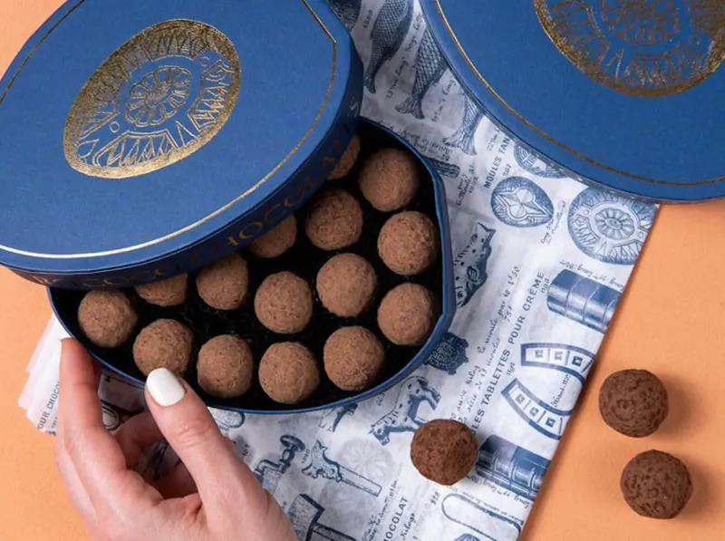 Open blue box with gold nautical design revealing round chocolate truffles, hand lifting lid, extras on blue-patterned cloth.