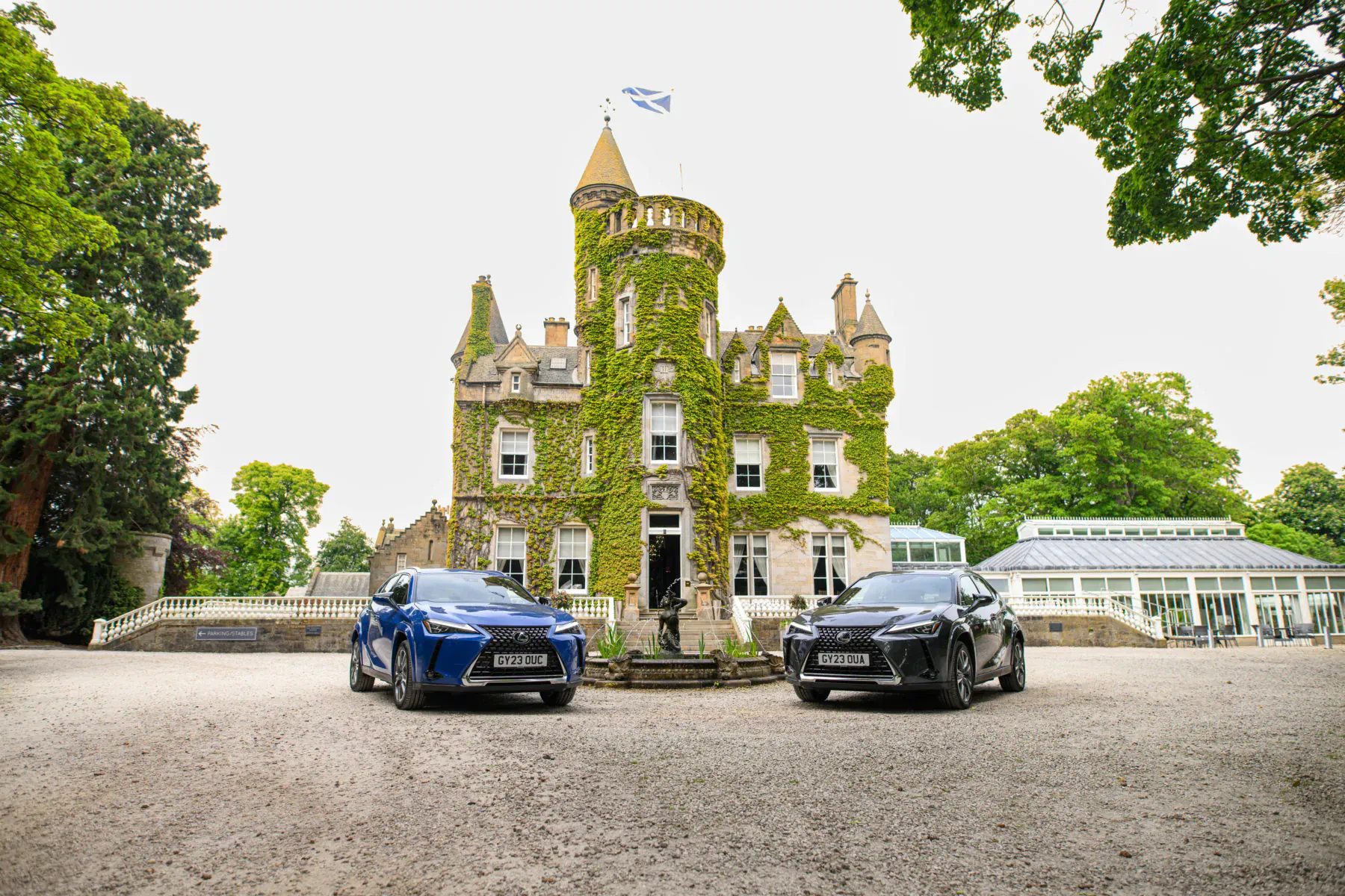 Ivy-covered turreted castle in Scotland with Scottish flag, blue and black Lexus cars parked in front, greenhouse nearby.
