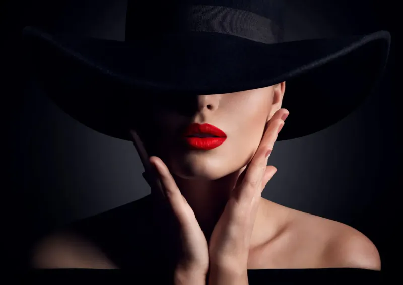 Close-up of woman's face shadowed by wide black hat, bold red lips, hands framing face, dark background