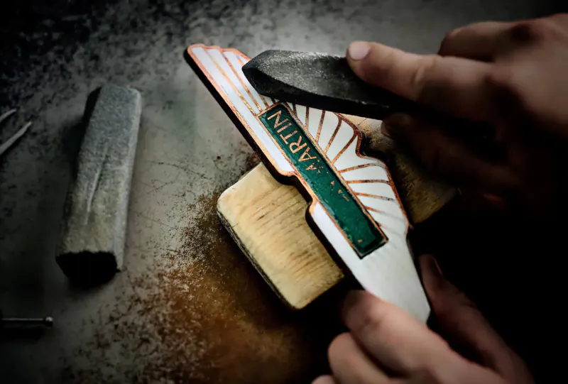 Hands sanding a wooden Aston Martin wings logo emblem with green 'Aston Martin' text on dusty surface.