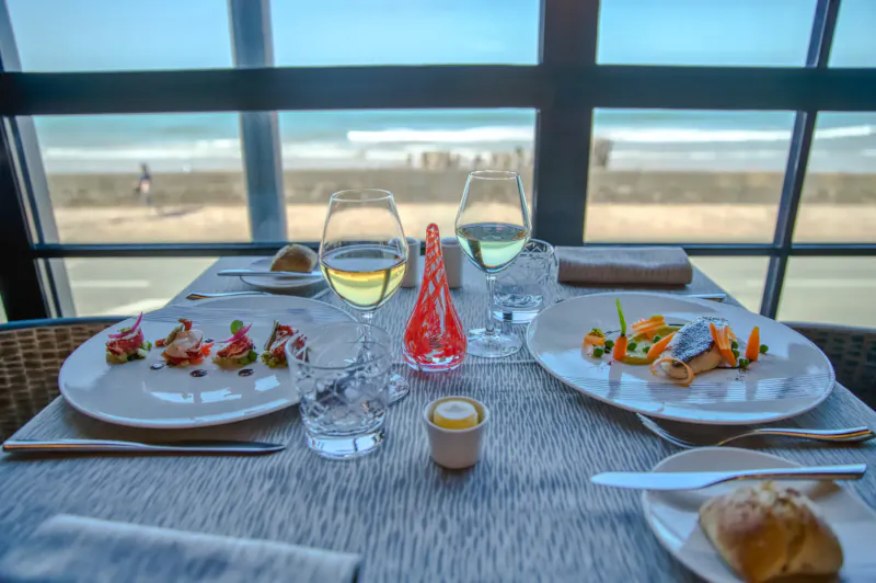 Elegant seaside restaurant table with seafood plates, white wine, red glass vase, and ocean view window at Saint-Malo hotel.