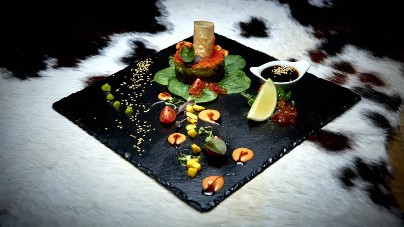 Stacked tuna tartare on square slate with greens, tomatoes, lemon, soy sauce, on fur backdrop