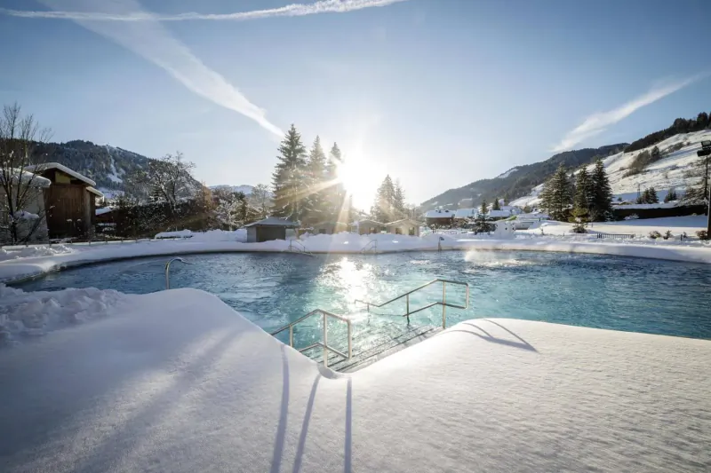 Snowy outdoor pool in Megève with turquoise water, surrounded by Alps, chalets, pine trees, and setting sun.