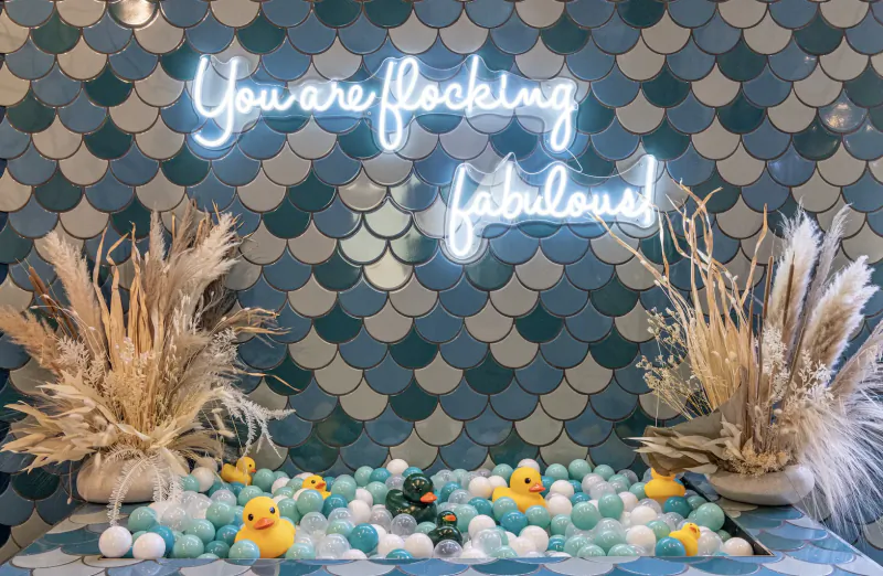 Neon 'You're Flocking Fabulous!' sign over blue ball pit with rubber ducks, pampas grass, and mermaid tile wall at Duck & Dry Galentine's event