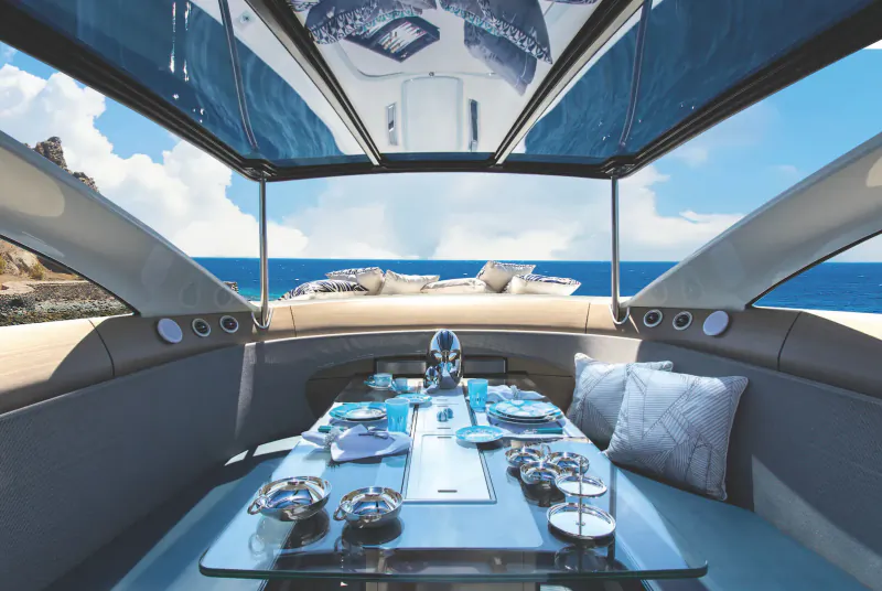 Luxury yacht cockpit with elegantly set dining table, blue sky, and sea view beyond rocky cliffs.