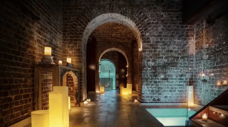 Tranquil brick arched corridor at Aire Ancient Baths, London, lit by glowing candles, leading to a pool.