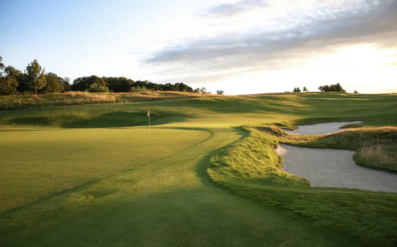 Aerial view of lush green golf course hole with bunkers, stream, and trees at sunset during tournament at The Grove.