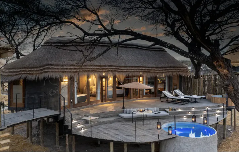 Luxury thatched Camp Kala lodge on elevated deck with blue hot tub, lounge chairs, umbrella, and acacia trees at sunset near Etosha.