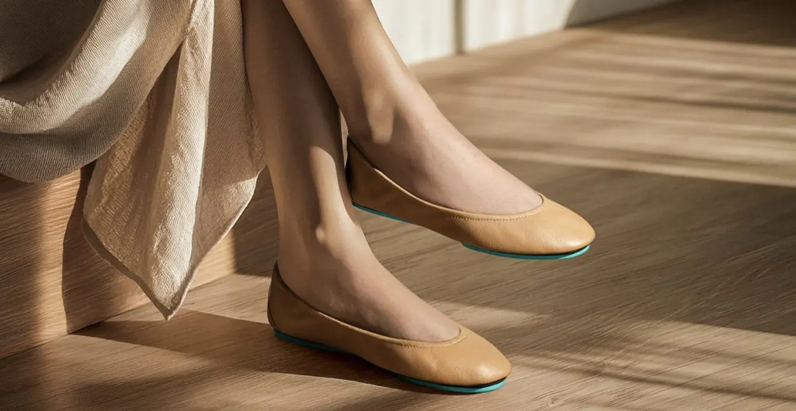 Woman's legs in beige Tieks ballet flats with turquoise soles, seated on wooden floor in sunlit room