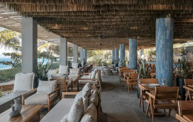 Open-air dining area at Amber Lombok with white lounges, wooden chairs, blue columns, palms, and ocean views.