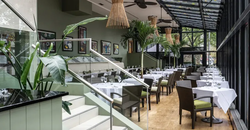 Elegant interior of The Bristol Gallery restaurant with white-clothed tables, green plants, rattan lamps, and glass atrium.