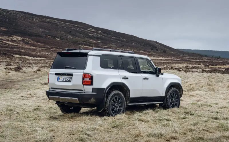 White Toyota Land Cruiser SUV, rear three-quarter view, parked on grassy hillside with 'FJ' plate, rugged terrain backdrop.
