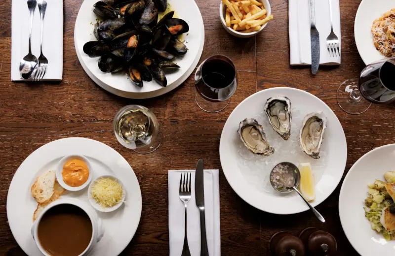 Top-down view of restaurant table with mussels, oysters, fries, soup, wine glasses, and cutlery on wood surface.