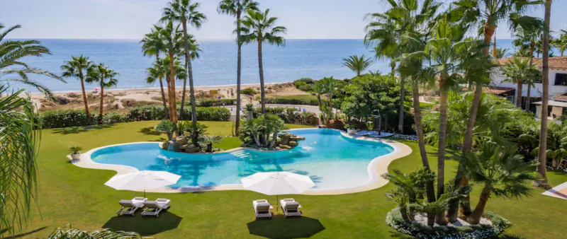 Aerial view of luxury villa resort in Marbella with lagoon pool, palm trees, loungers, and sea horizon.