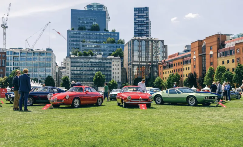 London Concours 2024: Classic V12 cars like red Ferrari and green supercar on green amid skyscrapers and crowd.