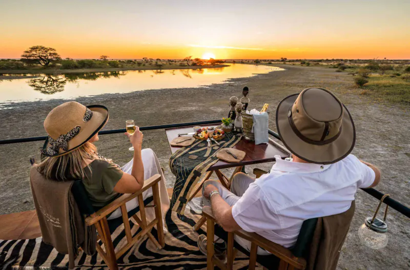 Couple in safari hats toasting wine at sunset table overlooking Etosha bushveld and lagoon from Onguma deck.