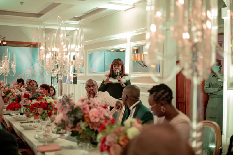 Woman speaking at podium at elegant Glenfiddich event with celebrities, chandeliers, and floral centerpieces in gallery.