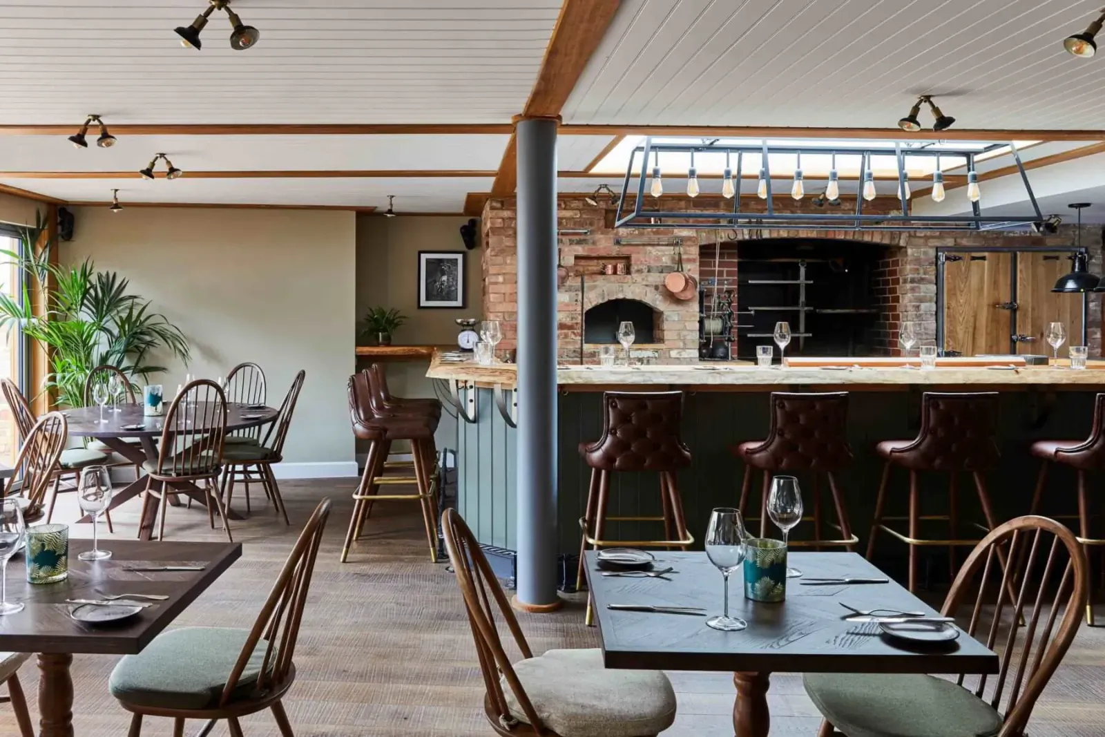 Cozy Hotel Indigo restaurant with brick wood-fired oven, wooden bar stools, tables set with wine glasses, and plants.