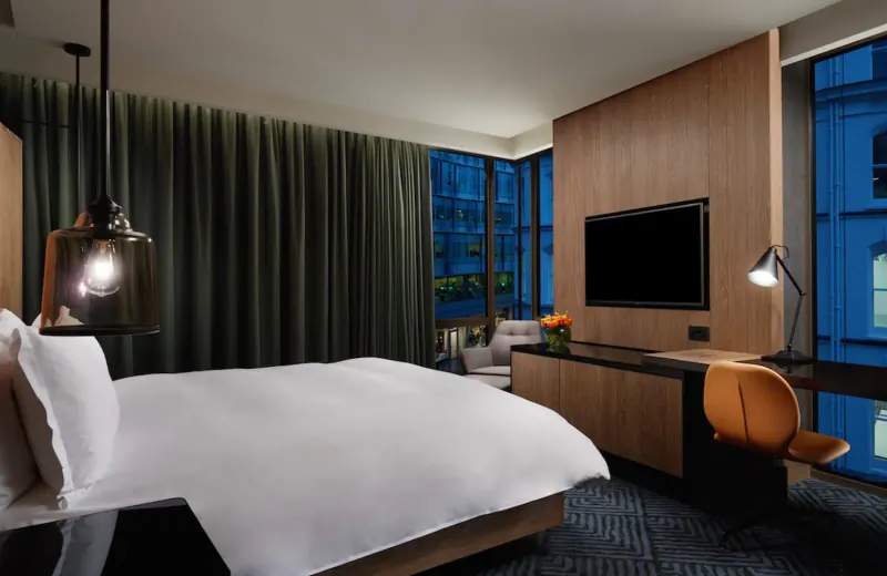 Modern hotel room at Hilton London Bankside with king bed, wood desk, orange chair, pendant lamps, and city night view.