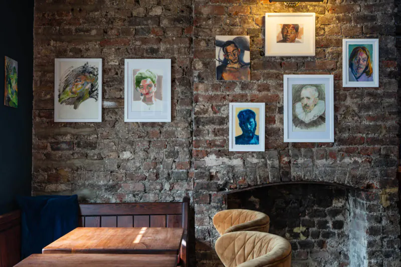 Gallery wall of colorful portrait paintings on brick above wooden bench and armchairs by fireplace.