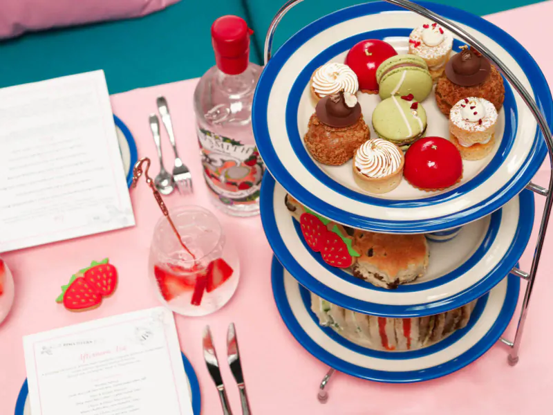 Three-tier blue and white cake stand with macarons, cupcakes, scones, sandwiches; gin bottle, pink drinks, strawberries on pink table.