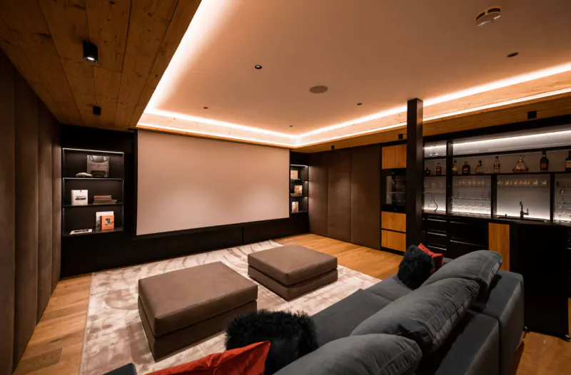 Luxurious home theater in Austrian chalet with projector screen, plush gray sofa, wood accents, and lit bar.