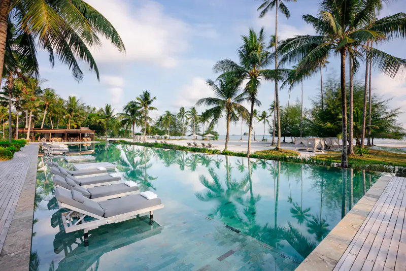 Infinity pool with white loungers at tropical beach resort, surrounded by palm trees and ocean under blue sky