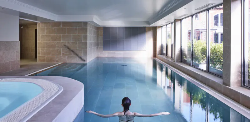 Woman with bun swims arms outstretched in indoor pool at Harper Norfolk resort, with jacuzzi and large windows.
