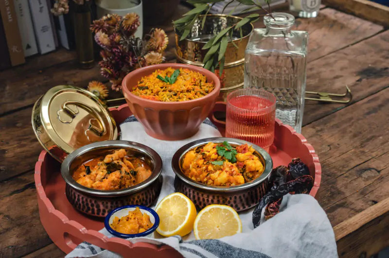 Indian dishes on wooden table: orange lentil curry, prawn masala in brass bowls, red drink, lemon, garnished with herbs.