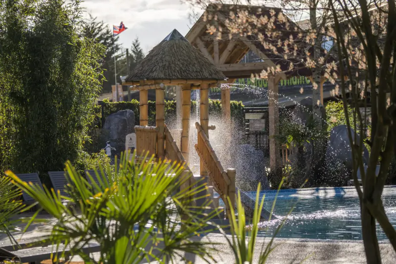 Wooden thatched pavilion with water cascade over stairs into blue pool, surrounded by lush greenery and palms at resort