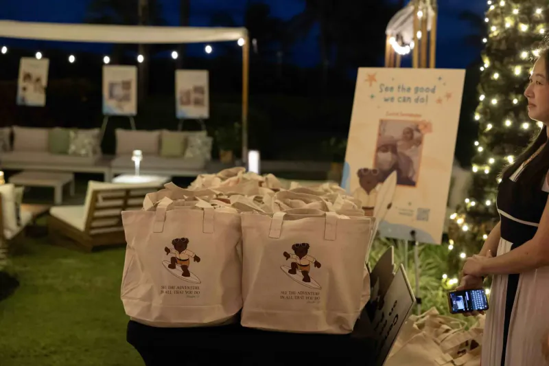 Woman holds phone by stacks of bear tote bags at outdoor evening charity event for childhood cancer society with 'We can do good' sign.
